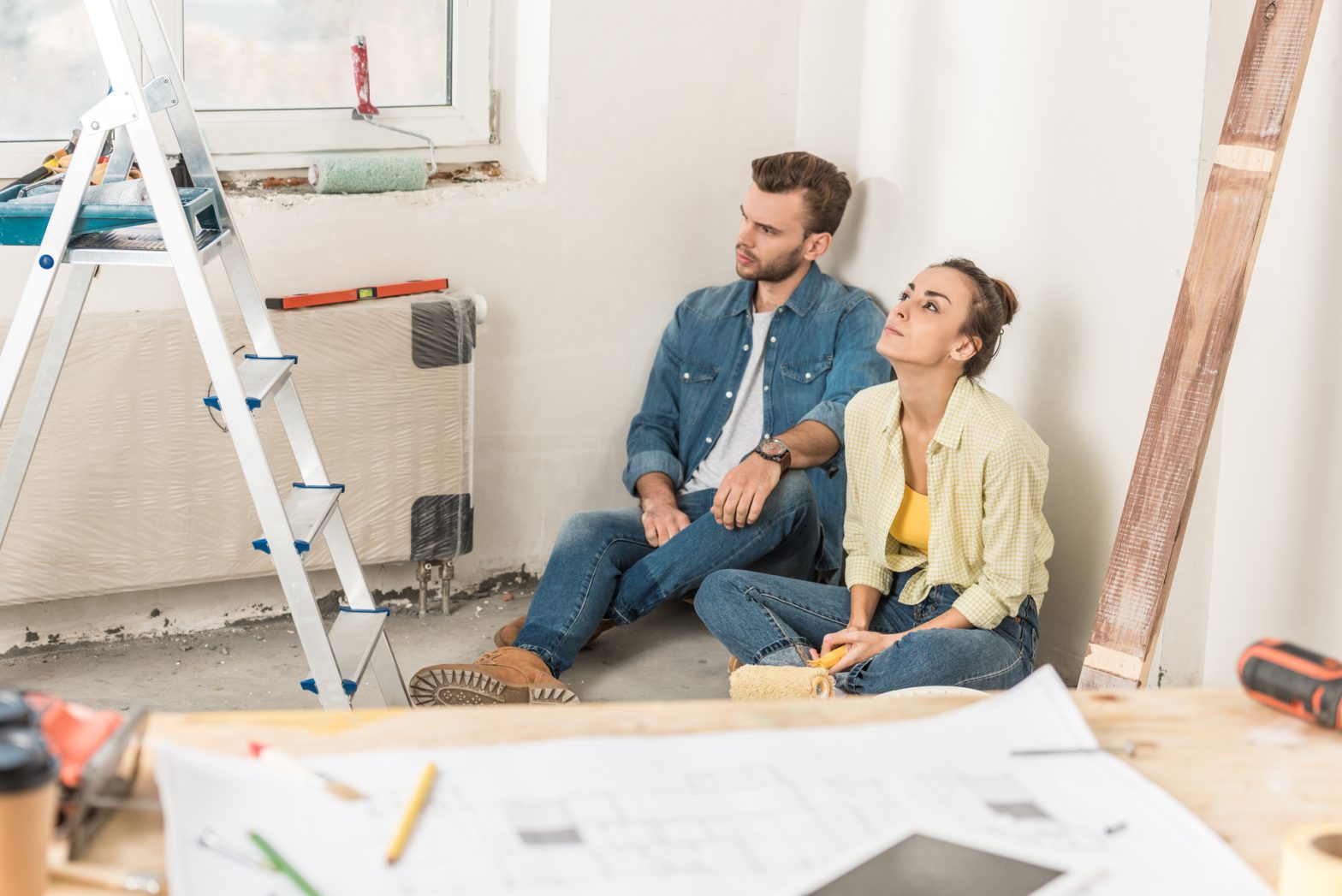 A young couple sits on the floor in their home, surrounded by paint and remodelling equipment