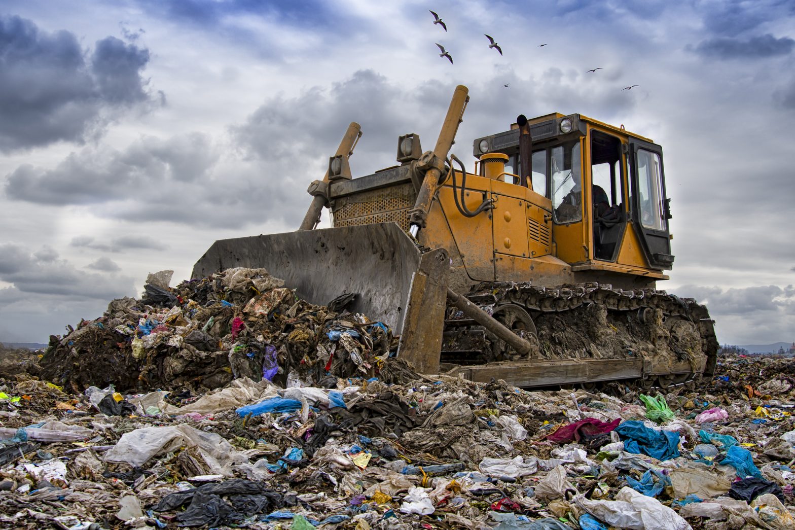 A yellow bulldozer atop a mound of trash, moving debris at a landfill.